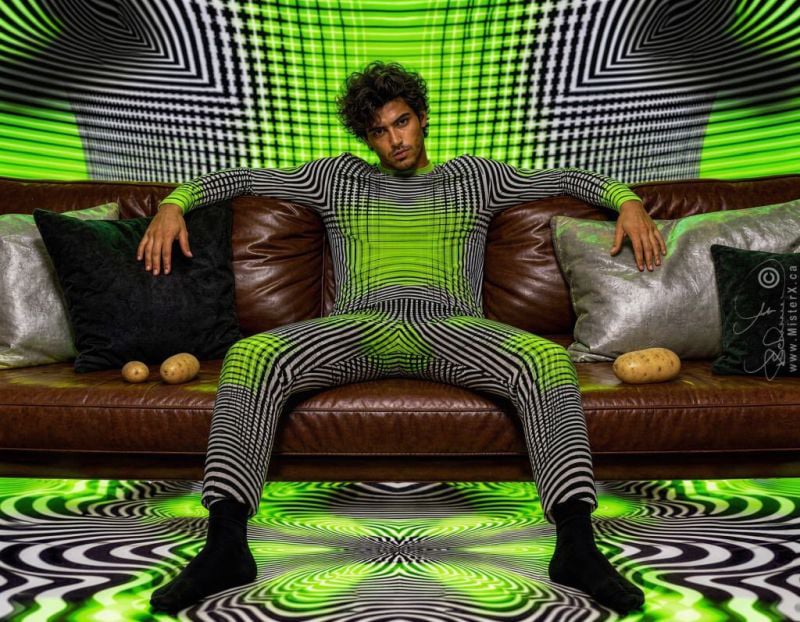 The image shows a man sitting on a brown leather sofa surrounded by intense, psychedelic patterns. He is wearing a tight-fitting, long-sleeve outfit covered in a black-and-white optical illusion pattern that is digitally overlaid with a vibrant neon green glow down the center. This same black, white, and neon green op-art pattern extends seamlessly onto the wall behind him.