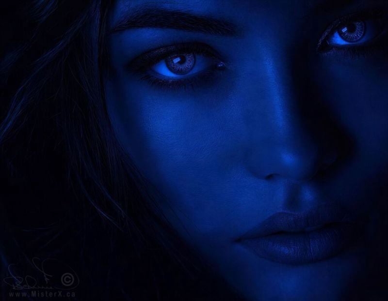 This moody digital artwork presents a close-up portrait of a woman's face, saturated entirely in deep monochromatic shades of midnight blue. Her intense, piercing eyes look directly at the viewer with an ethereal, patterned light in her irises that mirrors the texture of her skin, while her dark hair and the right side of her face dissolve softly into a pure black shadow background. The stark, low-key lighting accentuates the contours of her nose, cheekbones, and slightly parted lips, evoking a mysterious, nocturnal, or alien atmosphere that feels both serene and enigmatic.